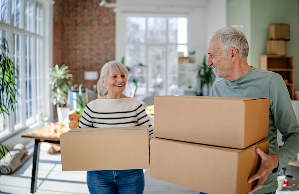 A retired couple carrying moving boxes into a bright new home as they prepare for winter living in a warmer location.