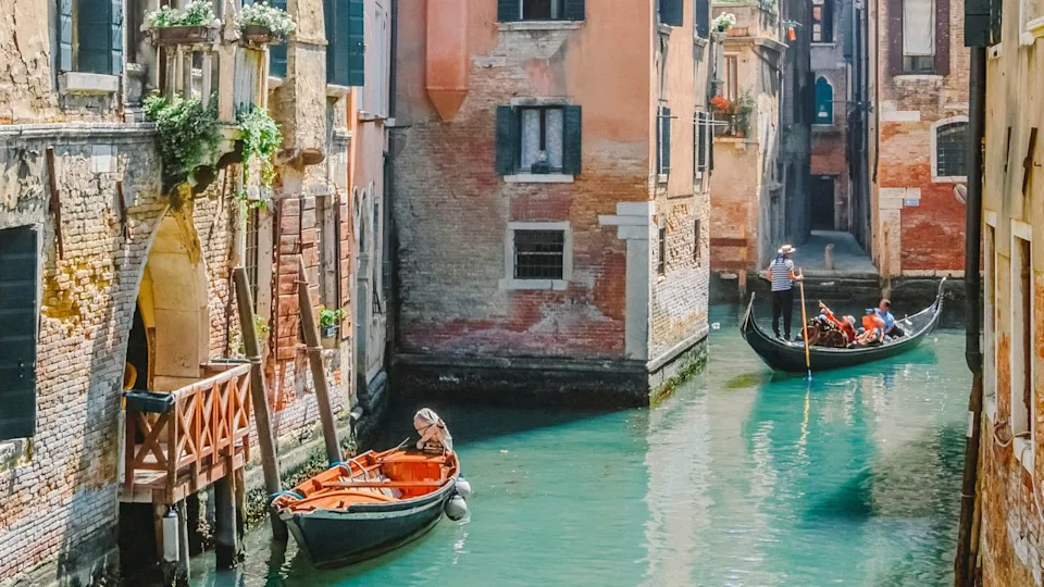 Gondolas on narrow canal and small boat tied next to old red brick house with wooden balcony on narrow canal in Venice, Italy.