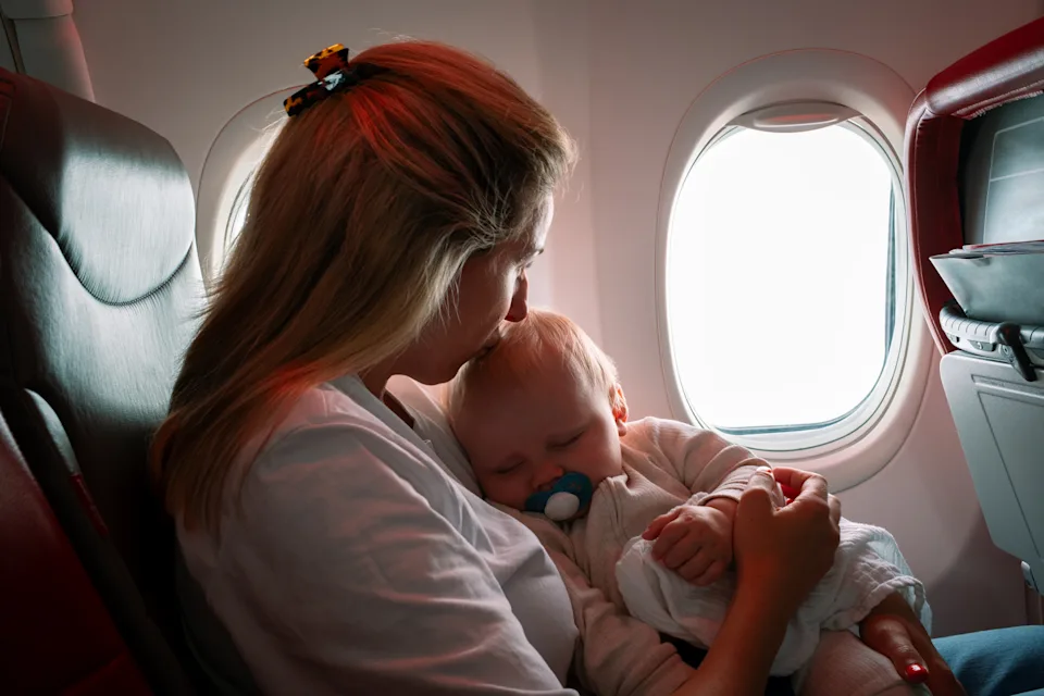 A waist-up shot of a mother holding her baby close while seated on an aeroplane. The baby boy has a pacifier in their mouth, sleeping peacefully in their parents’ arms during the flight. The mother is gently kissing her son's head. In Flight.