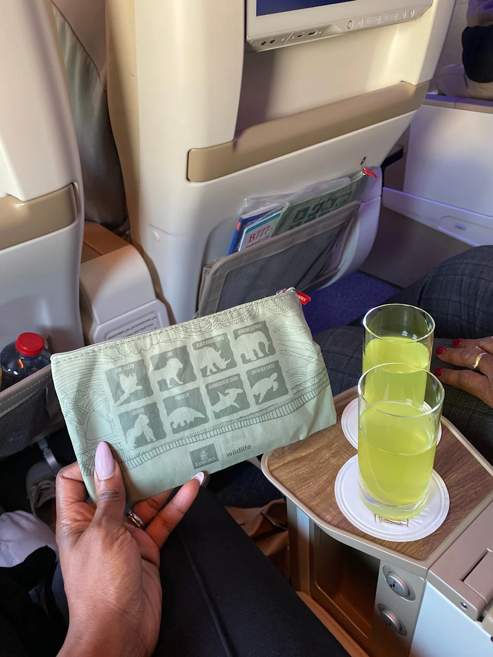 I am holding the Emirates Premium Economy amenity pouch while two glasses of welcome drinks rest on the wooden tray between the seats.