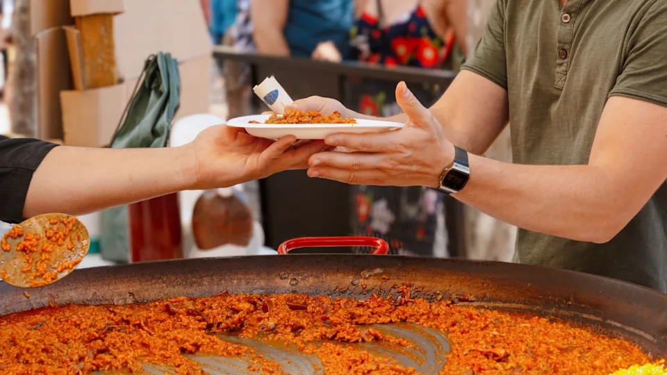 A steaming plate of paella is passed to a guest at a food stand with a large pan of rice during a street event in Valencia. Street food, Spanish paella