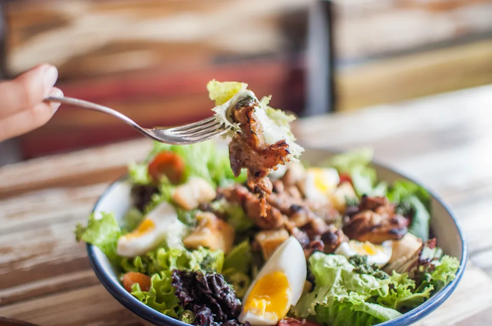 A fork holds a bite of salad with greens, chicken, and egg over a bowl, set on a wooden table, likely in a restaurant setting
