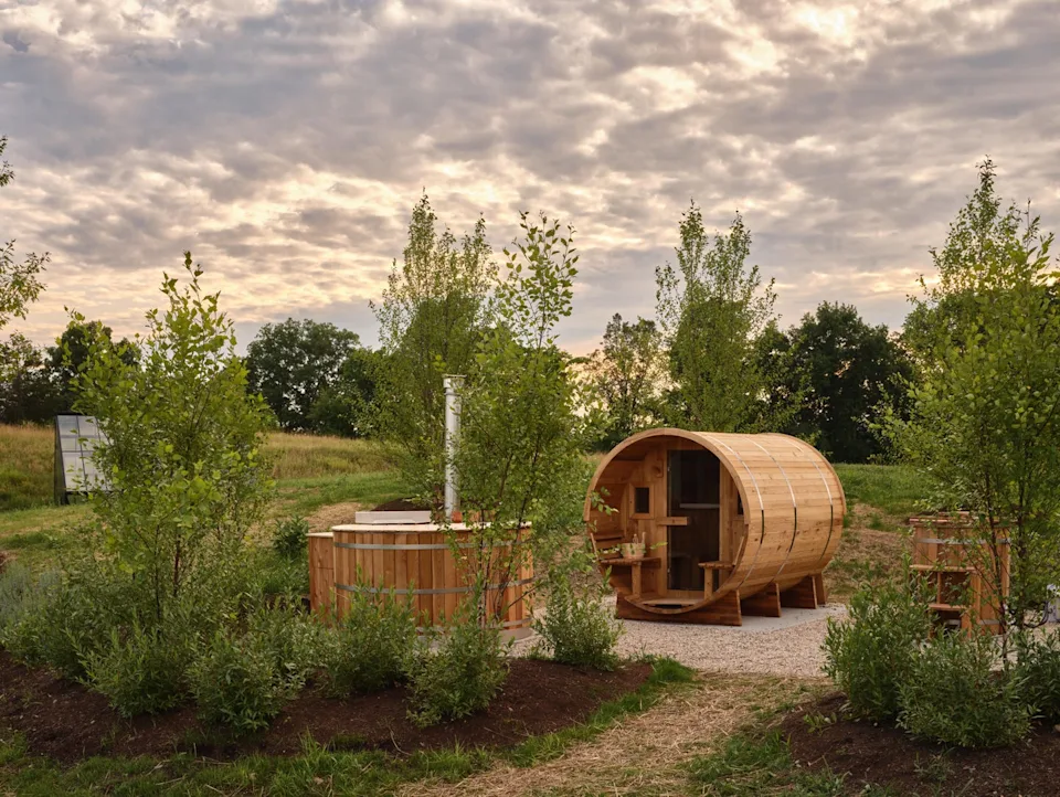 Outdoor wellness area with wooden barrel sauna and hot tub surrounded by greenery.