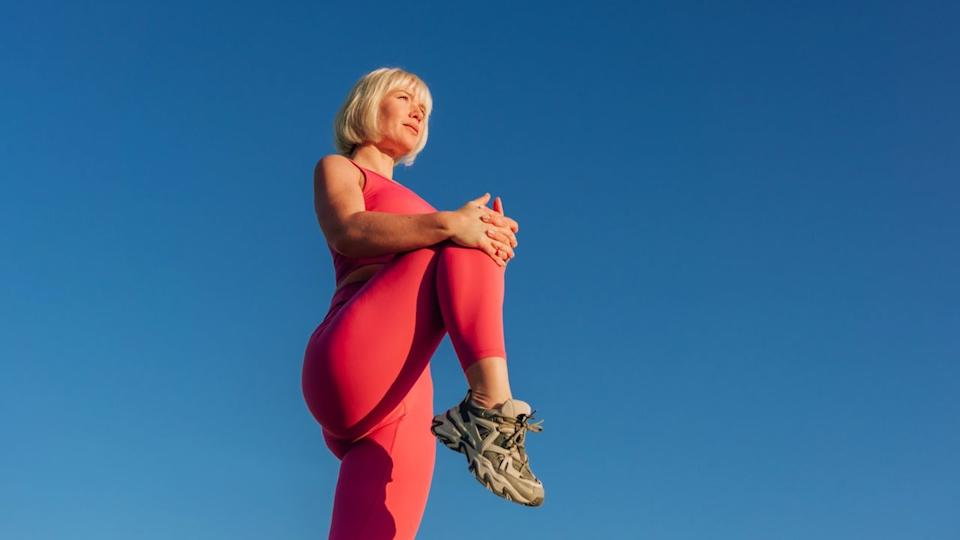  Blonde woman in pink workout gear against a blue sky backdrop holding her knee to her chest for a stretch. 