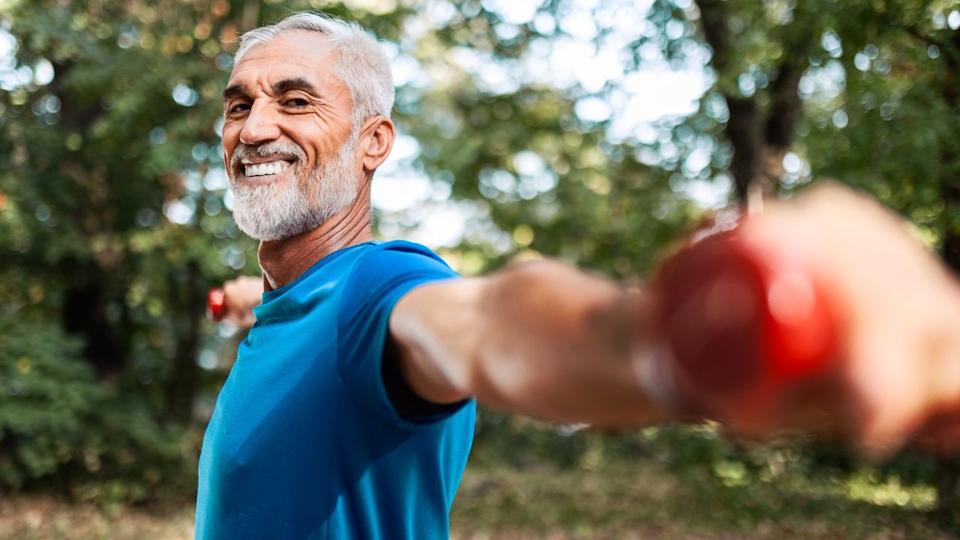  Older male in blue t-shirt holding dumbbells out at shoulder-height in a local park smiling. 