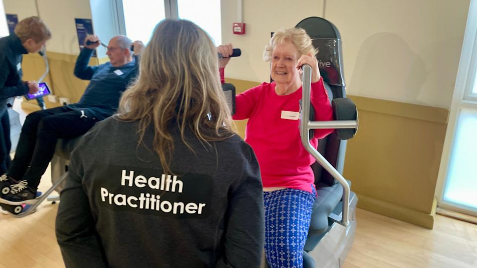A woman with back to us with the words 'Health Practitioner' on her sweat shirt and she is standing in front of an older woman who is sitting on a piece of rehabilitation equipment used to exercise her arms. 