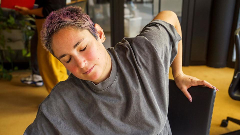 Person with cropped pink-dyed hair is leaning to the side in an office chair. They have their eyes closed and an expression of mild discomfort or fatigue on their face.