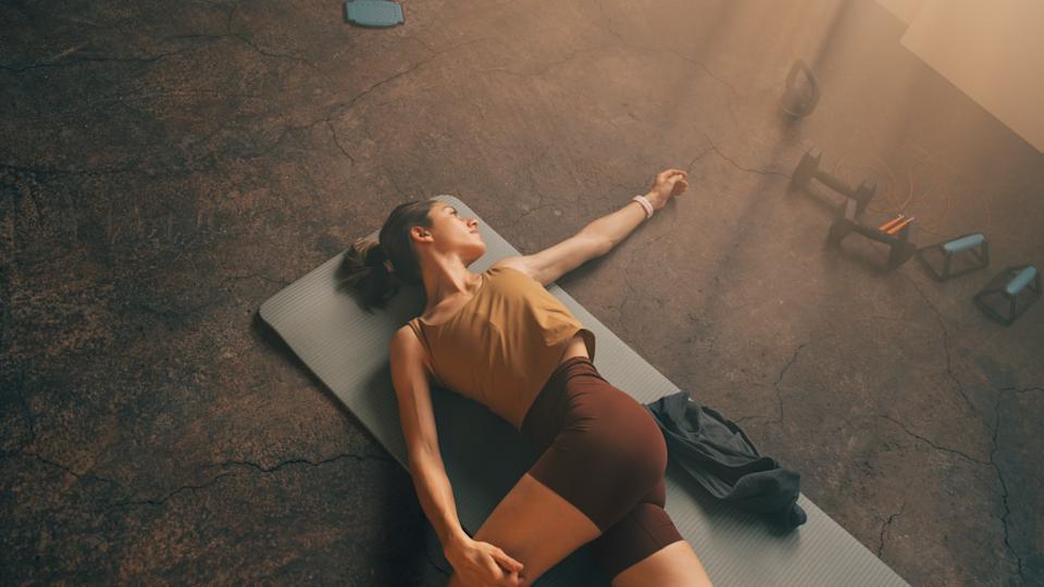 Woman lying on her back on yoga mat in studio with arm outstretched and leg over body for hip stretch 