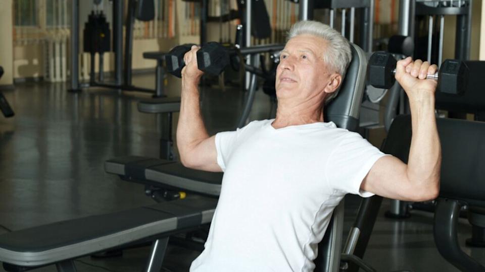 Elderly man in gym doing overhead press dumbbells, senior man exercising