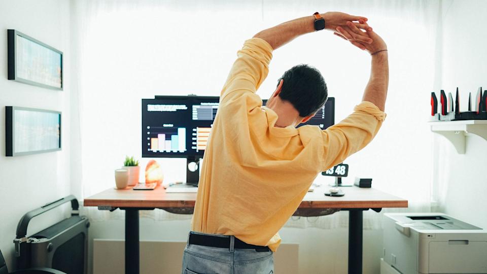 A man in a yellow shirt and jeans seen from the back. He has his arms above his head with his hands joined and is leaning to the side as if to stretch out his back