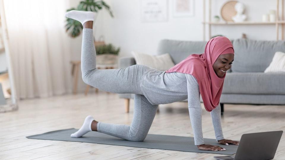  Woman dressed in light grey with a pink hijab on all fours with one leg kicking up behind her. she's on a grey exercise mat on wooden floor with a laptop on the floor in front of her. there's a grey sofa behind her. 