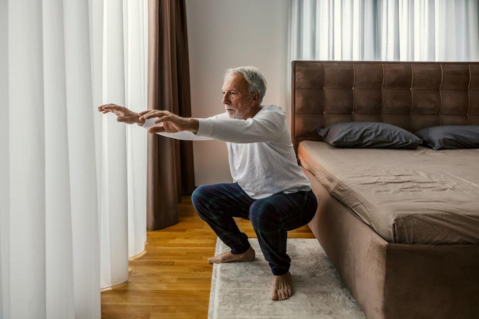  Man performing a squat looking off camera, wearing a white sleeved top and blue trousers in a bedroom setting on a cream rug and wooden floors. there's a brown bed next to him and white long net curtains in front. 
