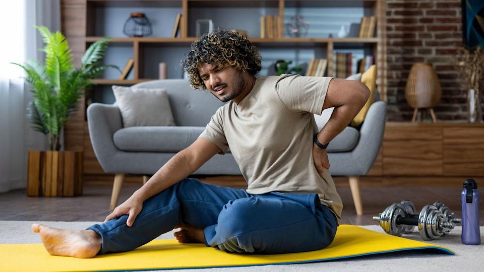  Man sits on yoga mat holding his back in pain. 