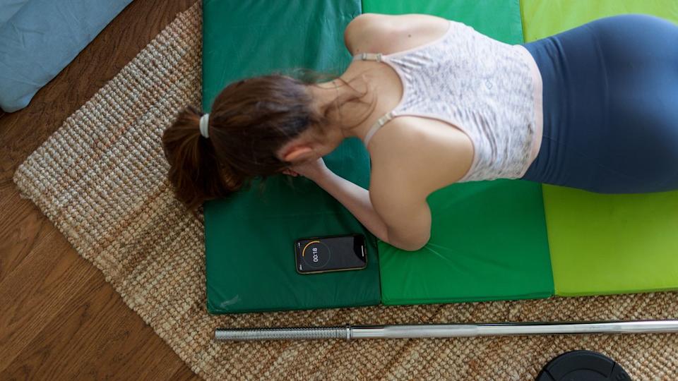 Woman seen from above holding a plank, a smartphone is next to displaying 18 seconds left on a timer.