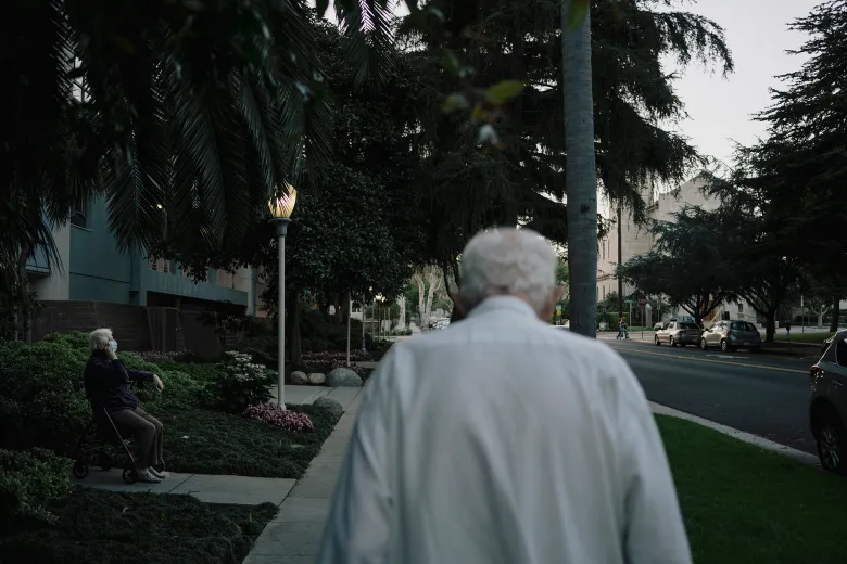 An older adult with white hair walks along a quiet, tree-lined sidewalk at dusk while another older adult wearing a face mask sits on a walker nearby, with parked cars and buildings lining the street.