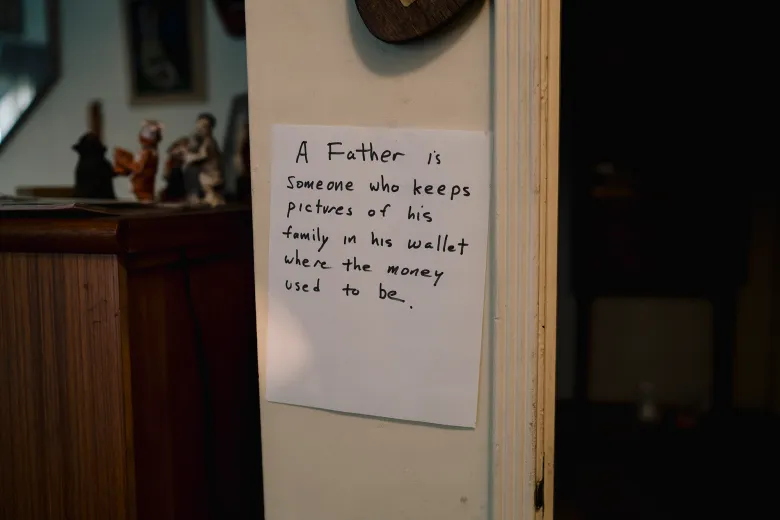 A handwritten note taped to a wall reads, “A Father is someone who keeps pictures of his family in his wallet where the money used to be,” beside a wooden cabinet topped with small figurines.
