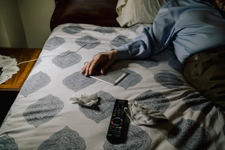 An older adult lies on a bed with a patterned gray-and-white cover, one hand resting on a tablet while a TV remote, a used tissue, and a face mask sit nearby.