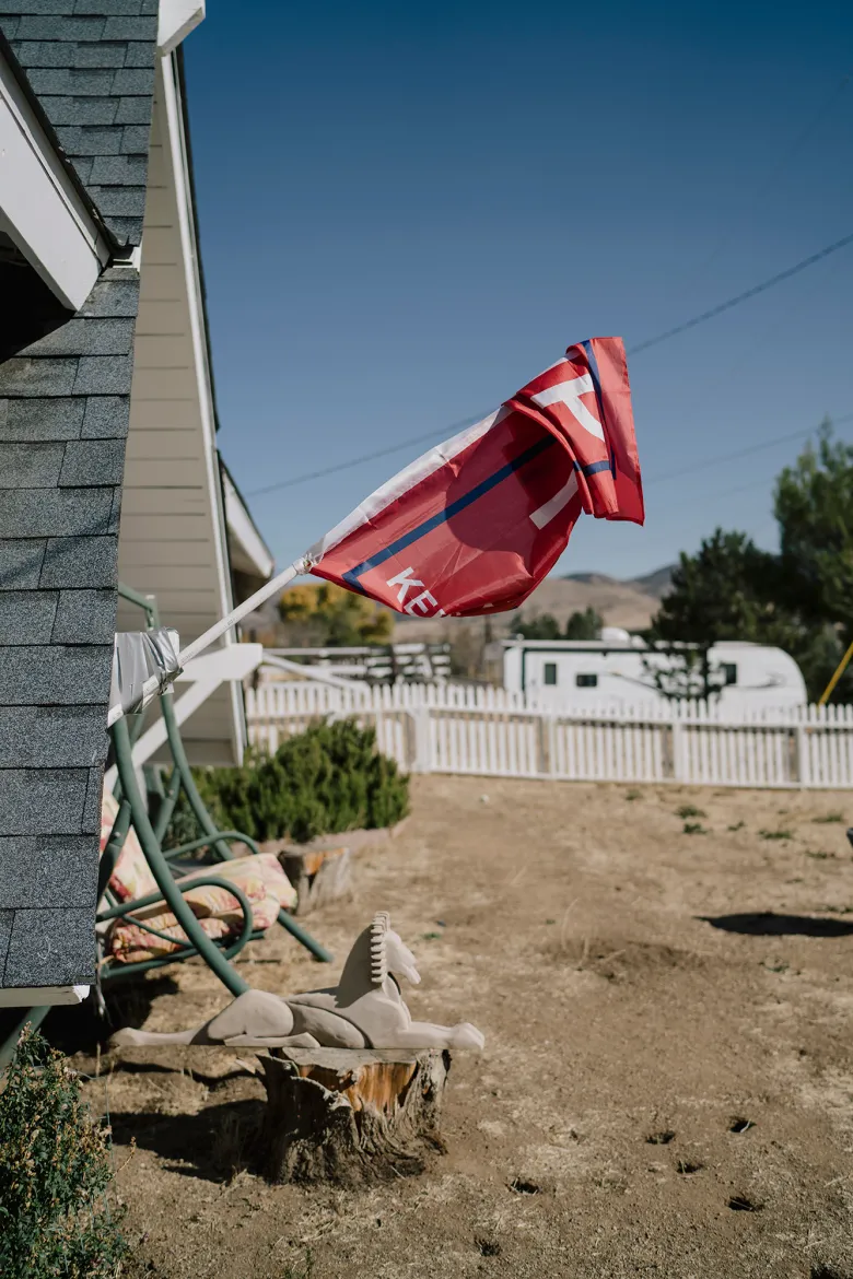 A red flag attached to the side of a house droops in still air above a dry yard with a white fence, a lawn swing, and a small decorative horse on a tree stump, with hills in the distance under a clear blue sky.