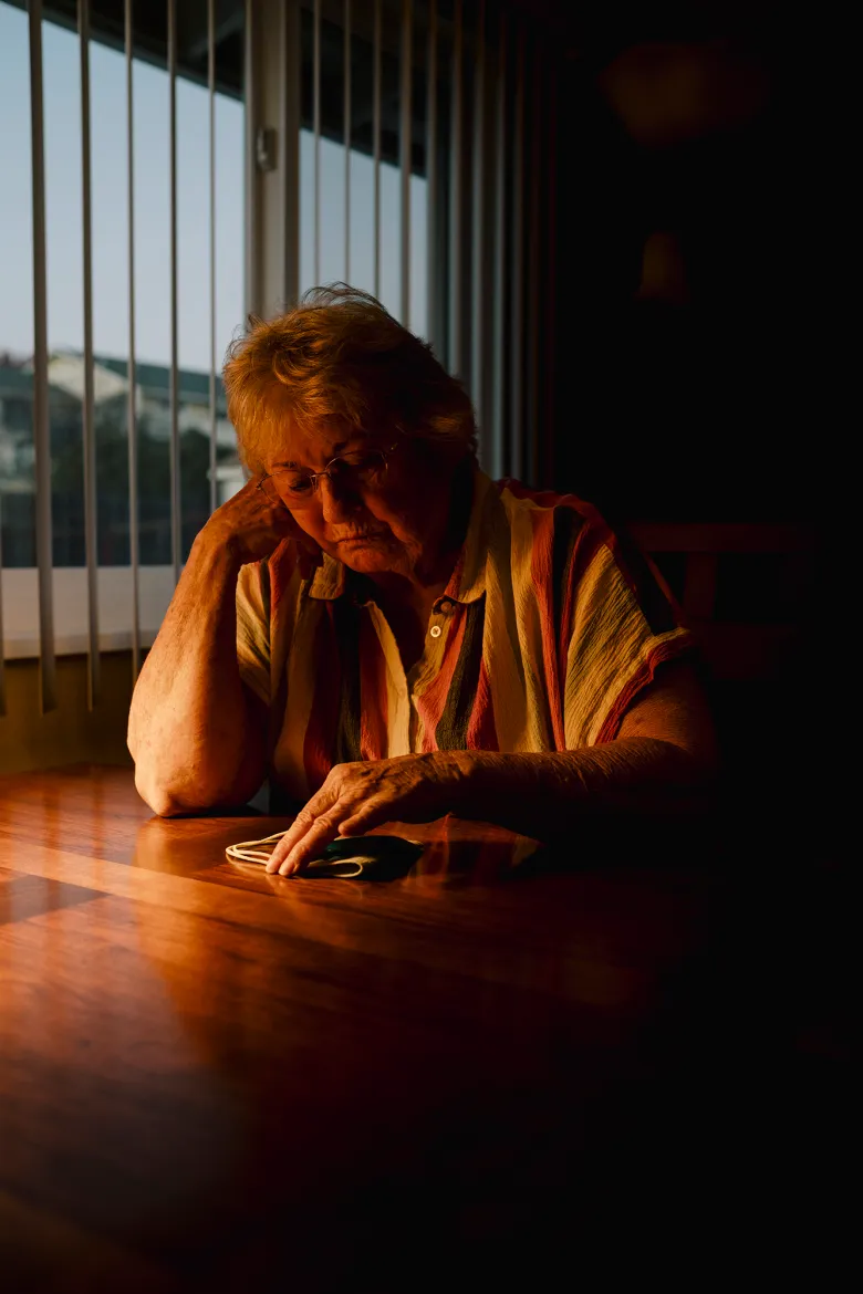 An older adult sits at a wooden table in warm evening light, resting their head on one hand while touching a face mask on the tabletop, with vertical blinds and a dim room behind them.