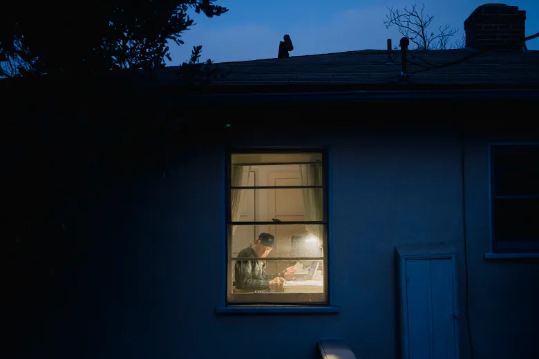 At dusk, an older adult sits at a desk inside a softly lit room, seen through a window from outside, reading papers while the exterior of the house remains in shadow.