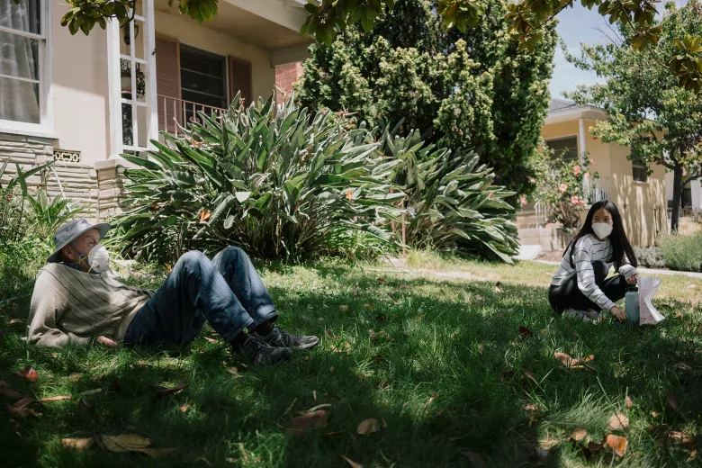 An older adult wearing a face mask reclines on the grass in a shaded yard while another masked person crouches several feet away holding a bag, with leafy plants and houses in the background.