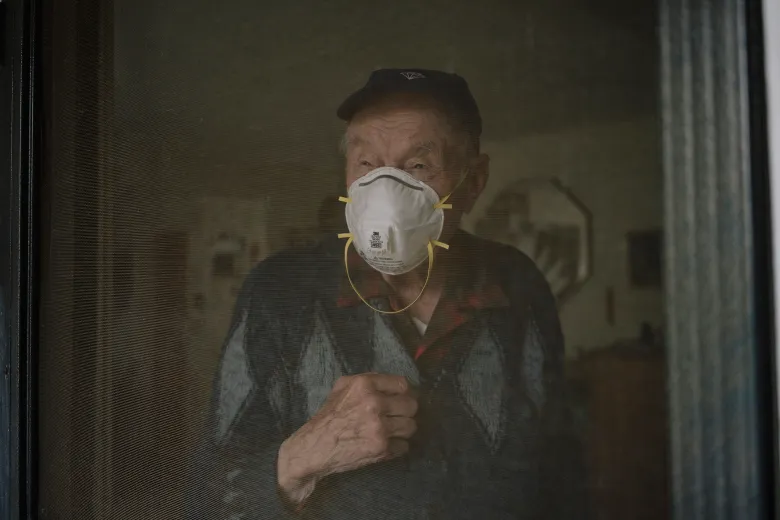 An older adult wearing a protective face mask and cap stands indoors behind a screen door, one hand resting near their chest as they look outward.