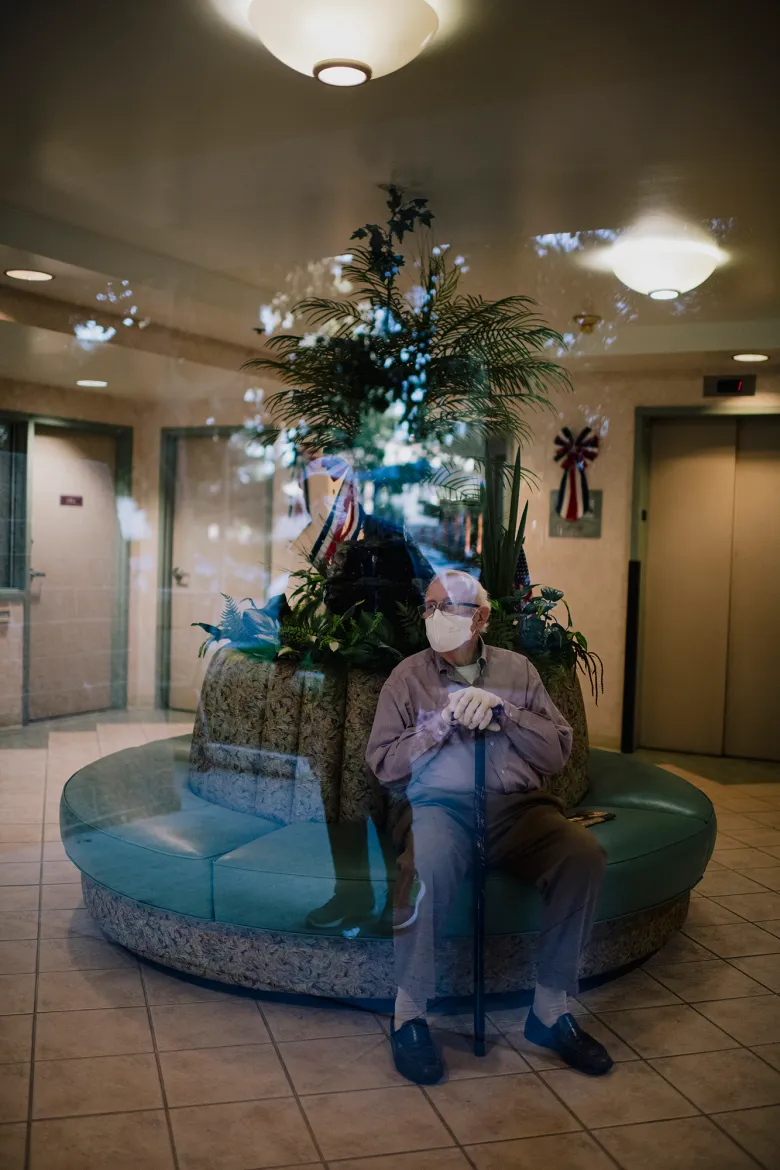 An elderly person wearing a face mask sits on a circular bench inside a lobby, holding a cane, with reflections of trees and sky layered over the scene through glass.