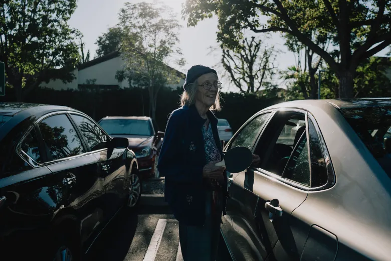 An older adult wearing a knit cap stands between parked cars in a sunlit lot, smiling while holding a small paddle and keys beside an open car door.