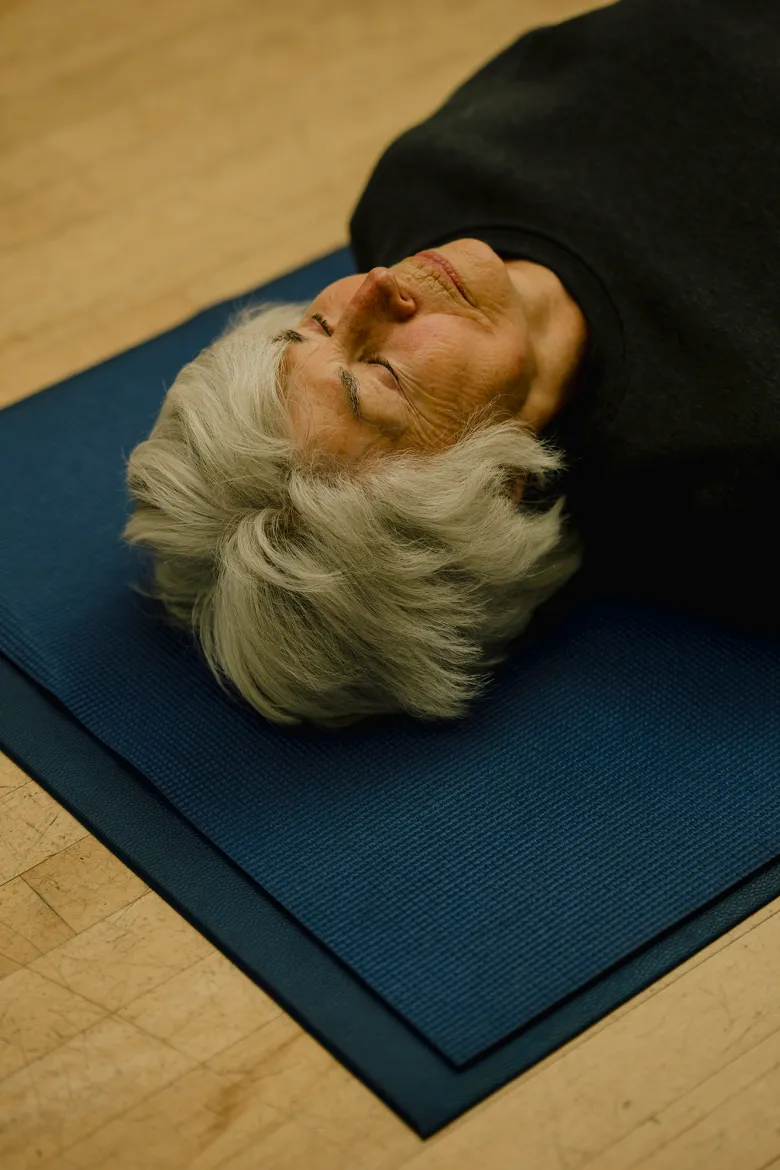 An older adult with short gray hair lies on their back on a blue exercise mat with eyes closed, appearing relaxed during a floor exercise or rest period.