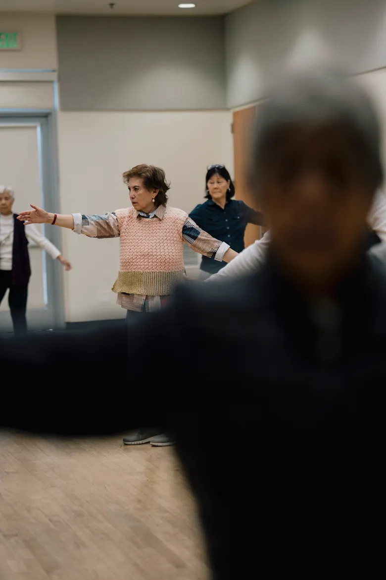 Several older adults stand spaced across a room with arms extended outward as if following a guided movement or exercise, while a blurred figure in the foreground partially obscures the view.
