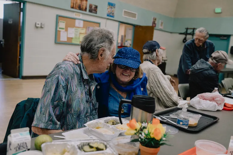 Two older adults sit closely together at a table in a community room, smiling and leaning in as one wraps an arm around the other’s shoulder, while several others sit nearby with trays of food and drinks.