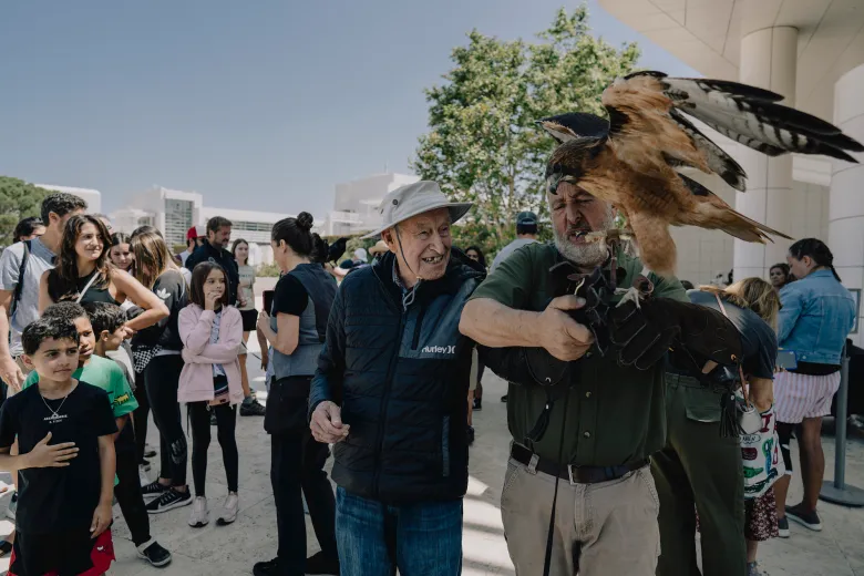 A handler wearing a glove steadies a large hawk as it flaps its wings near their face, while a smiling older adult in a sun hat watches closely. A crowd of adults and children stands nearby observing the demonstration outside a building.