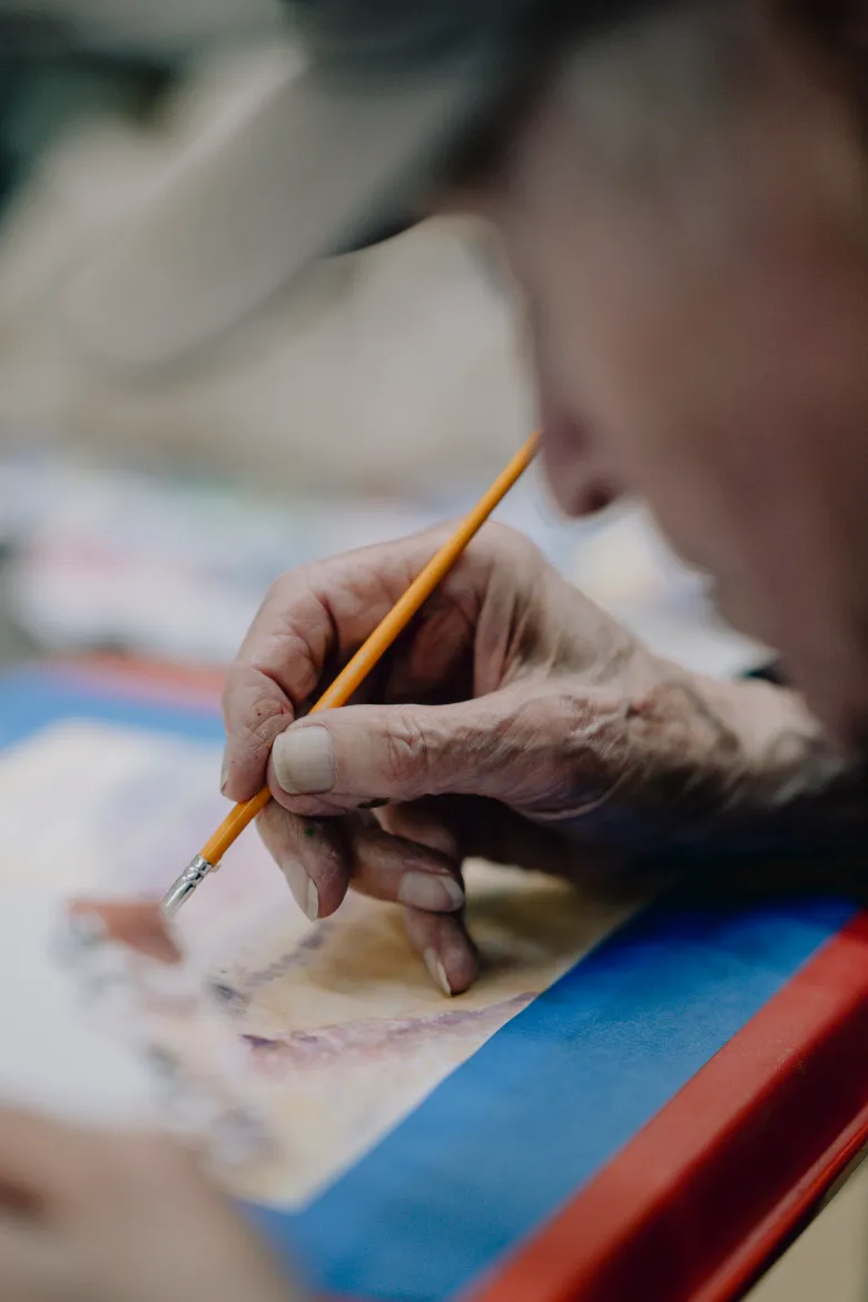 Close-up of an older adult’s hand holding a yellow pencil while carefully drawing or writing on paper, with their face softly out of focus in the foreground and a colorful tabletop beneath the page.