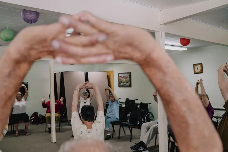 A group of older adults seated in a circle raise their arms overhead during a chair exercise class, while a pair of outstretched arms in the foreground frames the scene.