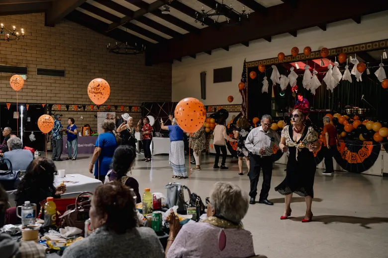 Older adults dance and socialize in a community hall decorated with orange balloons, ghost cutouts, and Halloween banners while a DJ plays music on a small stage.