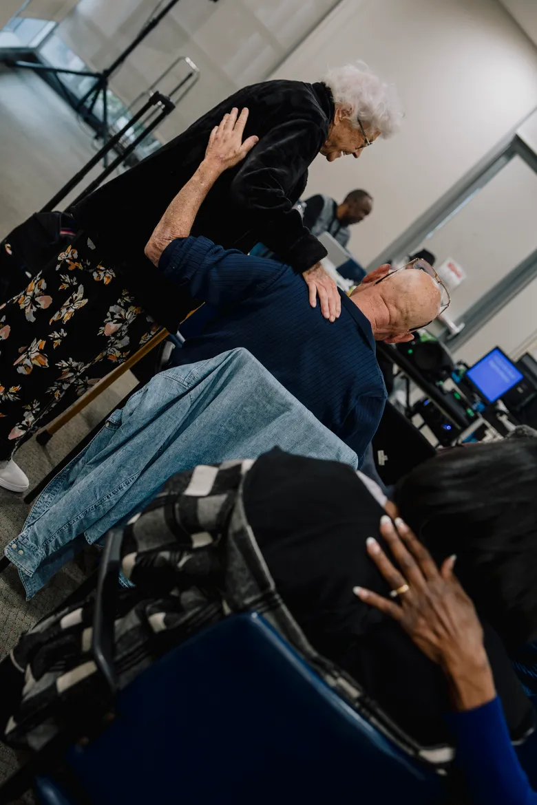 An older adult stands beside a seated participant and gently places a hand on their shoulder during a group activity, while others nearby mirror the gesture in a softly lit room.