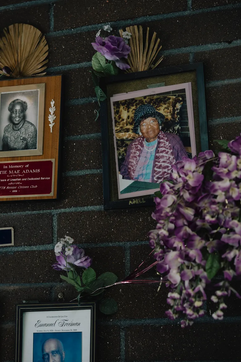 Framed memorial portraits and plaques hang on a brick wall, decorated with artificial purple flowers and small keepsakes honoring past community members.