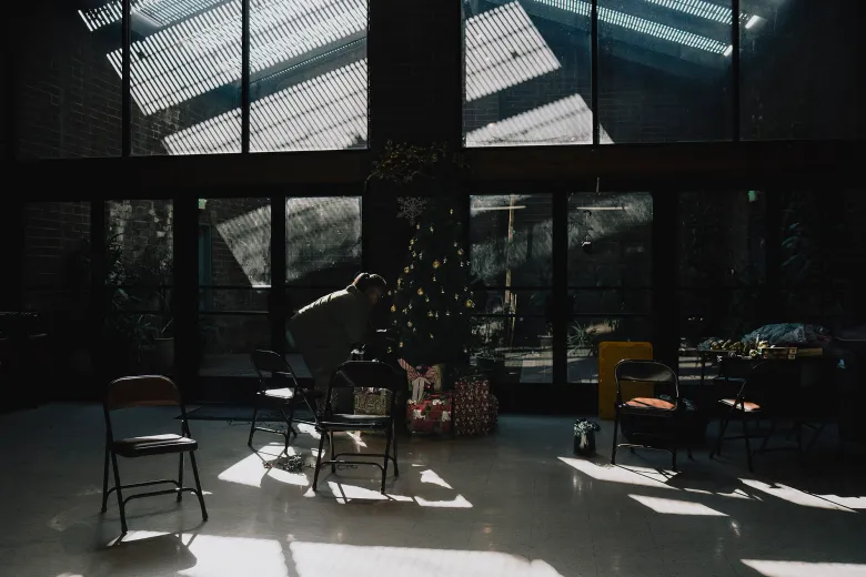 A person bends near a decorated Christmas tree surrounded by wrapped gifts in a dim community hall, where rows of empty folding chairs sit in shafts of sunlight streaming through tall windows.