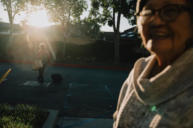 In warm sunset light, a smiling person in glasses and a scarf is blurred in the foreground while another person crosses a parking lot in the distance pulling a small rolling suitcase and carrying a tote bag.