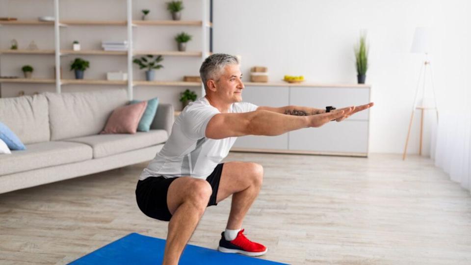 Stay home alone to workout. Fit mature man in sportswear doing squats in living room interior, exercising his legs