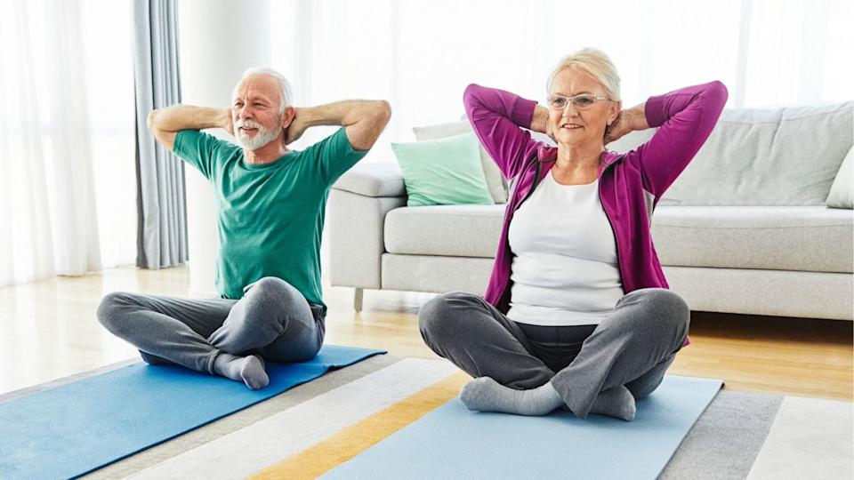  Senior couple sit on yoga mats doing a stretch, with hands behind their heads. 