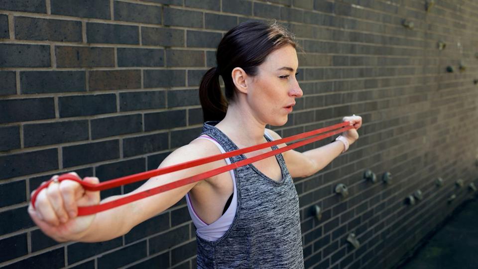  Woman in a grey vest standing sideways to the camera against a brick wall stretching a red long loop resistance band across her chest. 