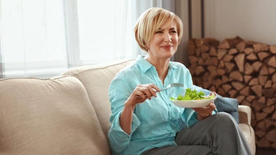 Mature Woman Eats Salad wearing In Casual Clothes With Healthy Food On Couch At Home