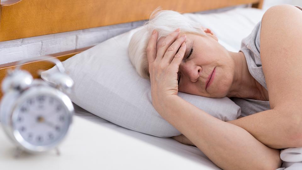 An older woman lies in bed with her head in her hands, struggling to sleep. At the front of the image is an out of focus alarm clock