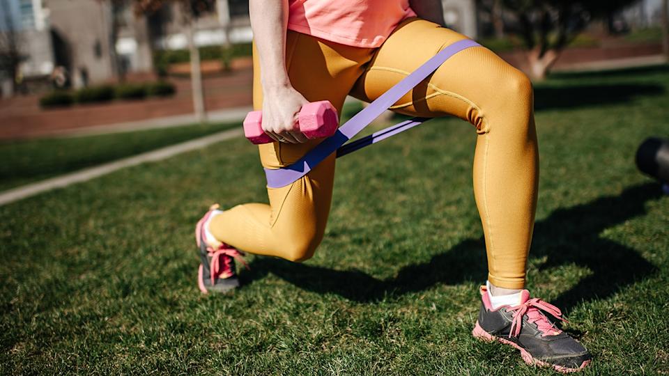 Woman performing a resistance band lunge with a dumbbell during an outdoor lower body workout on grass.