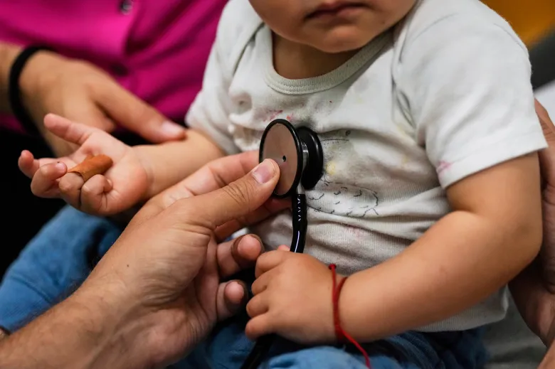 A healthcare worker holds a stethoscope against a young child's chest while the child sits on an adult's lap during a medical examination.