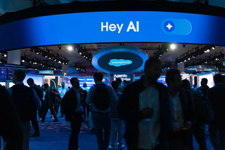 The silouhettes of people as they walk through a conference illuminated by blue LED signage. A main sign at the center of the room reads, "Hey AI."